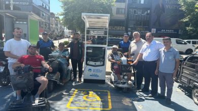 Görselde, engelli araçları için kurulan şarj istasyonunun açılışında çekilen bir toplu fotoğraf yer alıyor. Elektrikli tekerlekli sandalye ve mobilite scooterı kullanan bireyler ile yetkililer, şarj ünitesinin önünde bir araya gelerek poz veriyor. Zeminde engelli araçlarına ayrılmış şarj alanı işareti bulunurken, arka planda cadde üzerindeki iş yerleri ve binalar görülüyor.