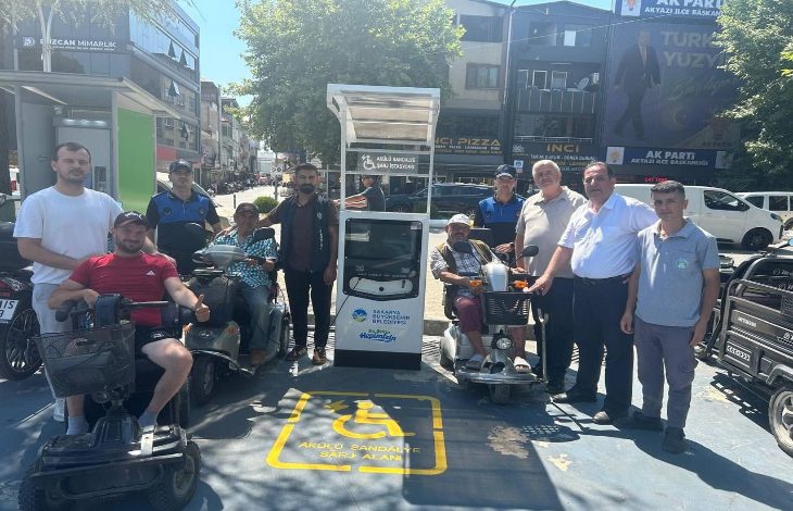 Görselde, engelli araçları için kurulan şarj istasyonunun açılışında çekilen bir toplu fotoğraf yer alıyor. Elektrikli tekerlekli sandalye ve mobilite scooterı kullanan bireyler ile yetkililer, şarj ünitesinin önünde bir araya gelerek poz veriyor. Zeminde engelli araçlarına ayrılmış şarj alanı işareti bulunurken, arka planda cadde üzerindeki iş yerleri ve binalar görülüyor.