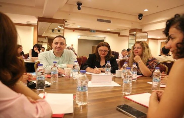 Görselde, toplantı salonunda yuvarlak bir masa etrafında oturan katılımcılar erişilebilirlik ve engelli hakları konusunda görüş alışverişinde bulunuyor. Masada su şişeleri, not defterleri ve telefonlar yer alırken, bir katılımcı not alıyor, diğerleri ise dikkatle tartışmayı takip ediyor.