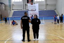 Görselde, kapalı spor salonunda bir golbol müsabakası öncesi veya sırasında hakem ile bir kadın orta sahada ayakta duruyor. Arka planda golbol oyuncuları, kale ve antrenman hazırlıkları görülürken, tribünler ve "T.C. Gençlik ve Spor Bakanlığı" yazılı pankart spor salonu ortamını yansıtıyor.