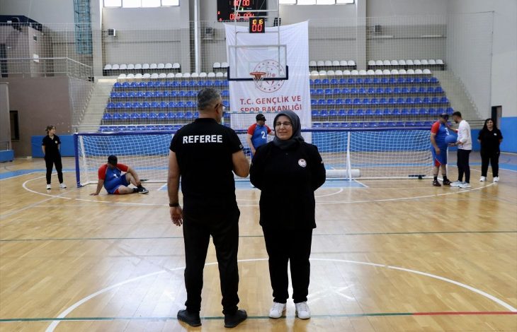 Görselde, kapalı spor salonunda bir golbol müsabakası öncesi veya sırasında hakem ile bir kadın orta sahada ayakta duruyor. Arka planda golbol oyuncuları, kale ve antrenman hazırlıkları görülürken, tribünler ve "T.C. Gençlik ve Spor Bakanlığı" yazılı pankart spor salonu ortamını yansıtıyor.