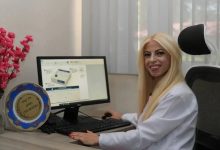 Görselde, beyaz önlük giyen bir sağlık çalışanı bilgisayar başında gülümseyerek kameraya poz veriyor. Bilgisayar ekranında dijital aşı kartına ait bir arayüz açıkken, masada klavye, fare ve üzerinde isim yazılı bir plaket yer alıyor. Görüntü, sağlık alanında geliştirilen dijital bir uygulamanın tanıtımını yansıtıyor.
