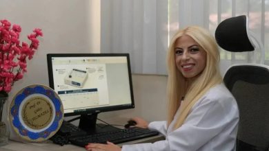 Görselde, beyaz önlük giyen bir sağlık çalışanı bilgisayar başında gülümseyerek kameraya poz veriyor. Bilgisayar ekranında dijital aşı kartına ait bir arayüz açıkken, masada klavye, fare ve üzerinde isim yazılı bir plaket yer alıyor. Görüntü, sağlık alanında geliştirilen dijital bir uygulamanın tanıtımını yansıtıyor.