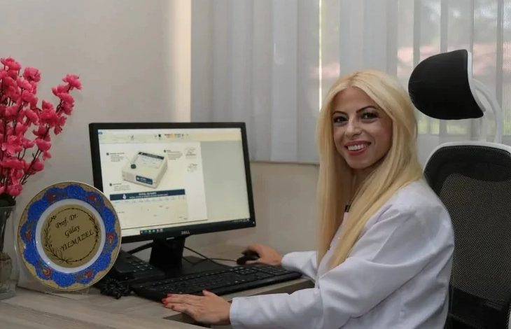 Görselde, beyaz önlük giyen bir sağlık çalışanı bilgisayar başında gülümseyerek kameraya poz veriyor. Bilgisayar ekranında dijital aşı kartına ait bir arayüz açıkken, masada klavye, fare ve üzerinde isim yazılı bir plaket yer alıyor. Görüntü, sağlık alanında geliştirilen dijital bir uygulamanın tanıtımını yansıtıyor.