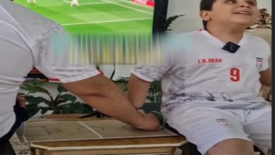 Görselde, İran milli takım forması giyen bir çocuk bir masanın kenarında otururken, yanında bulunan kişi kartondan yapılmış küçük bir futbol sahası maketi üzerinde elini hareket ettiriyor. Arka planda televizyonda futbol maçı yayınlanıyor. Sahne, çocuğun maçı dokunarak takip etmesine yardımcı olunan duygusal bir anı yansıtıyor.