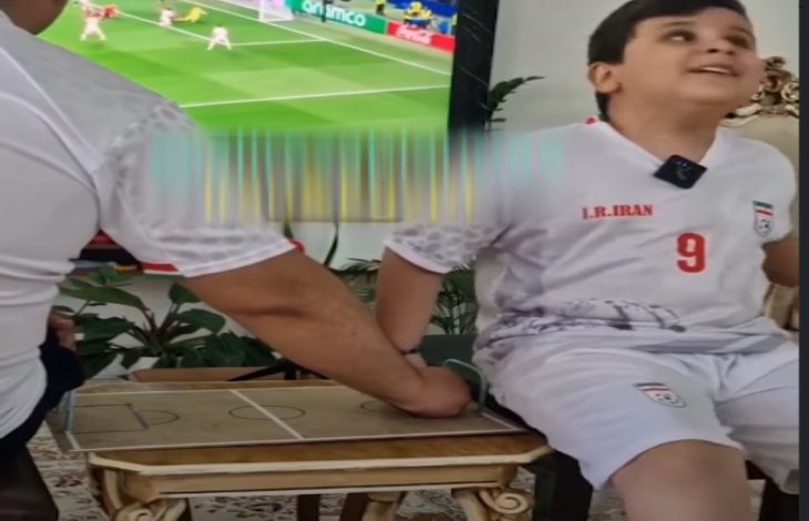 Görselde, İran milli takım forması giyen bir çocuk bir masanın kenarında otururken, yanında bulunan kişi kartondan yapılmış küçük bir futbol sahası maketi üzerinde elini hareket ettiriyor. Arka planda televizyonda futbol maçı yayınlanıyor. Sahne, çocuğun maçı dokunarak takip etmesine yardımcı olunan duygusal bir anı yansıtıyor.