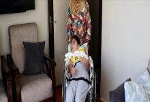 Bir ev ortamında, tekerlekli sandalyede oturan bir çocuk ve arkasında sandalyeyi yönlendiren bir yetişkin görülüyor. Odada koltuklar ve sade bir yaşam alanı yer alırken, görüntü bakım, destek ve günlük yaşam anını yansıtıyor.