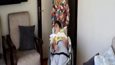 Bir ev ortamında, tekerlekli sandalyede oturan bir çocuk ve arkasında sandalyeyi yönlendiren bir yetişkin görülüyor. Odada koltuklar ve sade bir yaşam alanı yer alırken, görüntü bakım, destek ve günlük yaşam anını yansıtıyor.