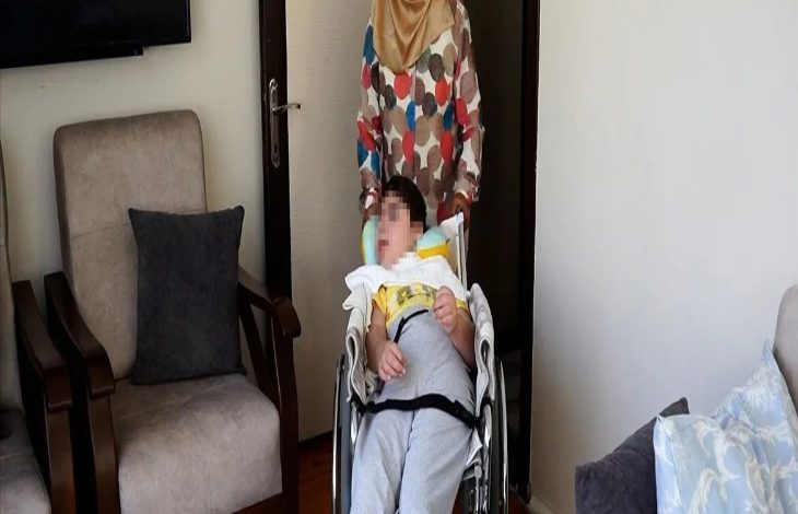 Bir ev ortamında, tekerlekli sandalyede oturan bir çocuk ve arkasında sandalyeyi yönlendiren bir yetişkin görülüyor. Odada koltuklar ve sade bir yaşam alanı yer alırken, görüntü bakım, destek ve günlük yaşam anını yansıtıyor.