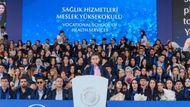 Görselde, bir üniversitenin mezuniyet töreninde kürsüde konuşma yapan bir öğrenci yer alıyor. Kürsünün arkasında mezuniyet cübbelerini giymiş çok sayıda öğrenci otururken, arka plandaki büyük ekranda Sağlık Hizmetleri Meslek Yüksekokulu mezuniyet törenine ait yazılar görülüyor. Atmosfer, kalabalık ve coşkulu bir mezuniyet törenini yansıtıyor.