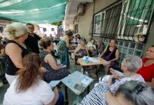Görselde, bir binanın açık alanında düzenlenen toplantıda çok sayıda kişi masa ve sandalyeler etrafında bir araya gelmiş durumda görülüyor. Katılımcılar sohbet ederken ve formları incelerken, masaların üzerinde su şişeleri ve belgeler yer alıyor. Açık hava ortamındaki buluşma, saha çalışması veya bilgilendirme etkinliği izlenimi veriyor.