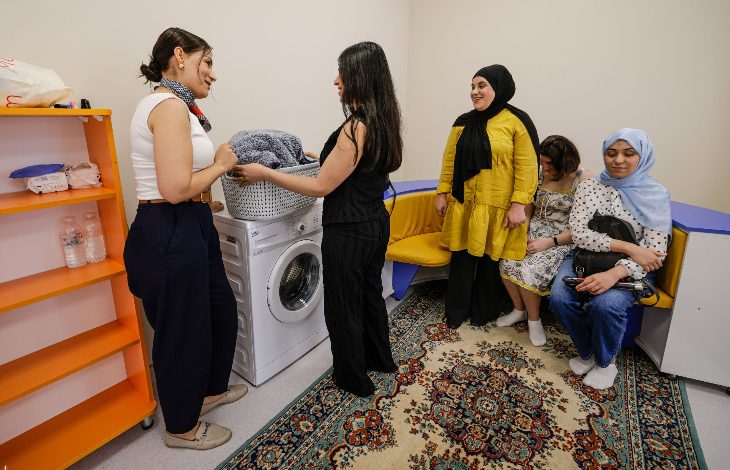 Görselde, engelli bireylerin günlük yaşam becerilerini geliştirmeye yönelik bir eğitim ortamı görülüyor. Ön planda iki kişi çamaşır makinesi başında uygulamalı çalışma yaparken, diğer katılımcılar oturma alanında etkinliği izliyor. Sınıf ortamında raflar, oturma grubu ve günlük yaşam eğitimine uygun ekipmanlar yer alıyor.