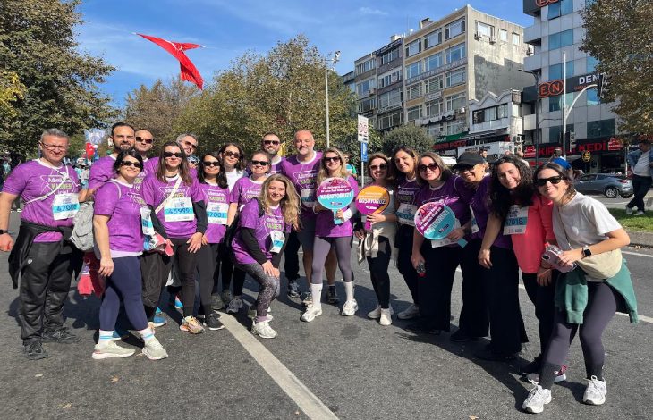Mor tişörtler giyen kalabalık bir grup, güneşli bir günde şehir caddesinde düzenlenen bir koşu etkinliği öncesinde gülümseyerek toplu hatıra fotoğrafı çektiriyor.