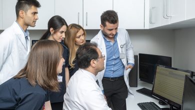 Prof. Dr. Selçuk Peker bir sandalyede oturuyor ve bilgisayar ekranına odaklanmış durumda. Etrafında ayakta duran diğer doktorlar ve tıp öğrencileri, ekranı dikkatle inceleyerek Peker’i takip ediyor. Ortam, beyaz dolapların ve tıbbi ekipmanların bulunduğu modern bir klinik çalışma alanı görünümünde. Ekip, birlikte bir vaka değerlendirmesi ya da eğitim amaçlı bir inceleme yapıyor izlenimi veriyor.