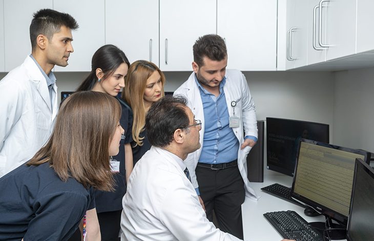 Prof. Dr. Selçuk Peker bir sandalyede oturuyor ve bilgisayar ekranına odaklanmış durumda. Etrafında ayakta duran diğer doktorlar ve tıp öğrencileri, ekranı dikkatle inceleyerek Peker’i takip ediyor. Ortam, beyaz dolapların ve tıbbi ekipmanların bulunduğu modern bir klinik çalışma alanı görünümünde. Ekip, birlikte bir vaka değerlendirmesi ya da eğitim amaçlı bir inceleme yapıyor izlenimi veriyor.