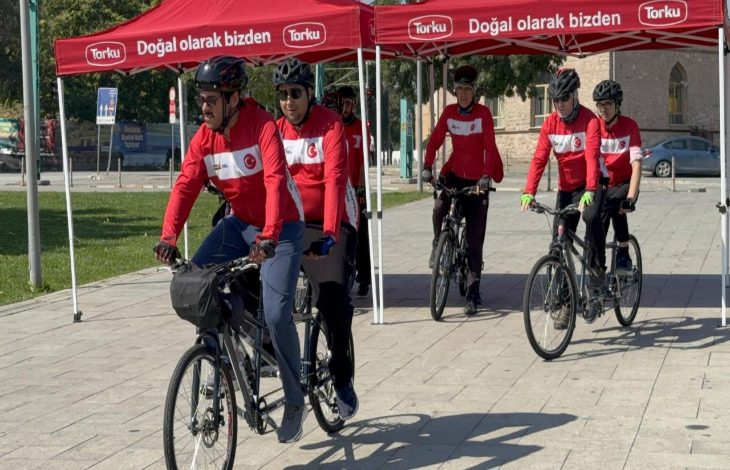 Görselde, kırmızı-beyaz formalar ve kasklar giyen tandem bisiklet sürücüleri, kırmızı çadırların önünden toplu halde hareket ediyor. Önde görme engelli sporcular ve onlara eşlik eden pilot sürücüler yer alırken, etkinlik açık havada gerçekleştiriliyor.