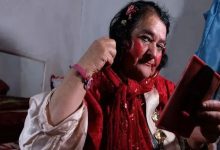 Görselde, kırmızı şal ve belirgin kırmızı makyajıyla dikkat çeken yaşlı bir kişi, elindeki aynaya bakarak saçını düzeltirken görülüyor. İç mekânda çekilen fotoğrafta, beyaz gömlek giyen kişinin bileğinde pembe bir bileklik bulunuyor ve arka planda sade bir oda ile yatak yer alıyor.