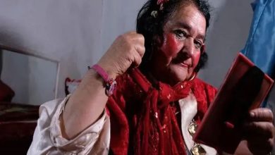 Görselde, kırmızı şal ve belirgin kırmızı makyajıyla dikkat çeken yaşlı bir kişi, elindeki aynaya bakarak saçını düzeltirken görülüyor. İç mekânda çekilen fotoğrafta, beyaz gömlek giyen kişinin bileğinde pembe bir bileklik bulunuyor ve arka planda sade bir oda ile yatak yer alıyor.