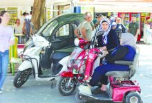 Görselde, kaldırım kenarında park edilmiş bir akülü engelli aracı ile üç tekerlekli elektrikli motosikletin yanında sohbet eden iki kadın görülüyor. Arka planda yaya trafiğinin yoğun olduğu çarşı alanı, dükkânlar ve vatandaşlar yer alırken, günlük yaşam içinde erişilebilir ulaşım araçları dikkat çekiyor.