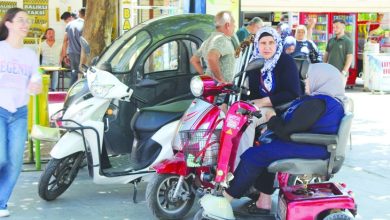 Görselde, kaldırım kenarında park edilmiş bir akülü engelli aracı ile üç tekerlekli elektrikli motosikletin yanında sohbet eden iki kadın görülüyor. Arka planda yaya trafiğinin yoğun olduğu çarşı alanı, dükkânlar ve vatandaşlar yer alırken, günlük yaşam içinde erişilebilir ulaşım araçları dikkat çekiyor.