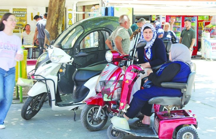 Görselde, kaldırım kenarında park edilmiş bir akülü engelli aracı ile üç tekerlekli elektrikli motosikletin yanında sohbet eden iki kadın görülüyor. Arka planda yaya trafiğinin yoğun olduğu çarşı alanı, dükkânlar ve vatandaşlar yer alırken, günlük yaşam içinde erişilebilir ulaşım araçları dikkat çekiyor.