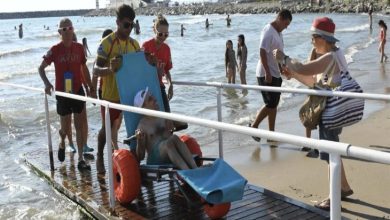 Görselde, özel tekerlekli deniz sandalyesi kullanan bedensel engelli bir birey, görevlilerin yardımıyla rampadan denize ulaştırılıyor. Plajda bulunan diğer tatilciler ve erişilebilirlik düzenlemeleri, engelsiz plaj uygulamasının kullanımını gösteriyor.