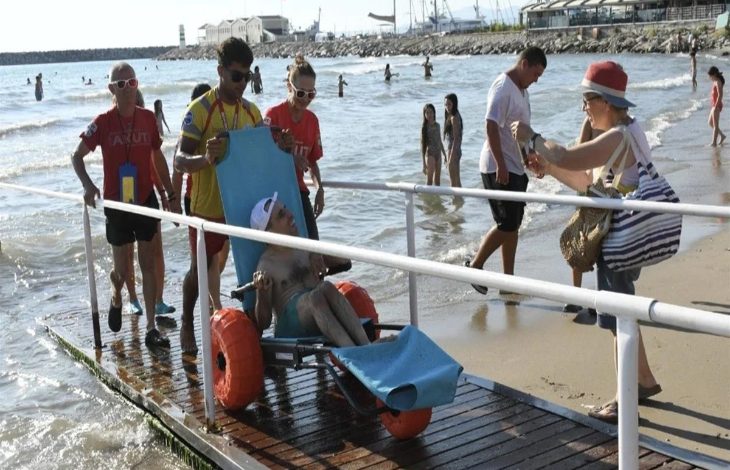 Görselde, özel tekerlekli deniz sandalyesi kullanan bedensel engelli bir birey, görevlilerin yardımıyla rampadan denize ulaştırılıyor. Plajda bulunan diğer tatilciler ve erişilebilirlik düzenlemeleri, engelsiz plaj uygulamasının kullanımını gösteriyor.