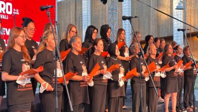 Sahnede siyah tişörtleri giymiş, ellerinde turuncu dosyalar tutan farklı yaş gruplarından kadınlar, mikrofonlar önünde koro düzeninde performans sergiliyor.
