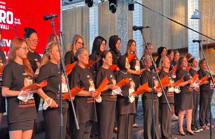 Sahnede siyah tişörtleri giymiş, ellerinde turuncu dosyalar tutan farklı yaş gruplarından kadınlar, mikrofonlar önünde koro düzeninde performans sergiliyor.