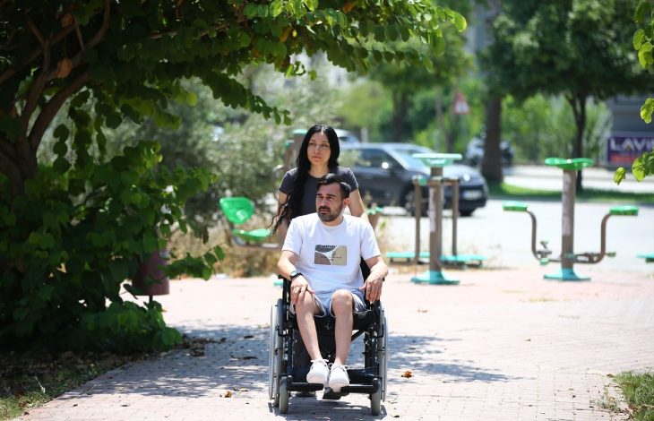 Mert Mutluay tekerlekli sandalyede oturuyor ve eşi sokakta aracını sürüyor.