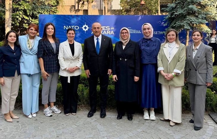 Taş döşeli açık alanda, NATO ve SETA logolarının yer aldığı büyük mavi bir afişin önünde dokuz kişilik bir grup fotoğrafı çekilmiş. Grubun ortasında, koyu renk takım elbisesiyle eski Dışişleri Bakanı Mevlüt Çavuşoğlu yer alıyor. Sağdan ikinci sırada açık renk kıyafetiyle SERÇEV Genel Sekreteri Edibe Sinem Ersoy bulunuyor. Her iki isim de kameraya dönük, resmi bir etkinlik atmosferi içinde poz veriyor. Arka planda yeşillikler ve ağaçlar görülüyor.