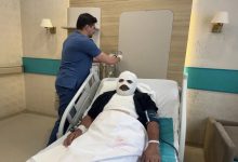 Görselde, hastane odasında tedavi gören bir hasta hastane yatağında uzanıyor. Hastanın başı ve vücudunun büyük bir bölümü bandajlarla sarılıyken, yanında bir sağlık çalışanı serum ekipmanıyla ilgileniyor. Oda, tıbbi ekipmanlar ve hasta yatağıyla donatılmış bir hastane ortamını yansıtıyor.