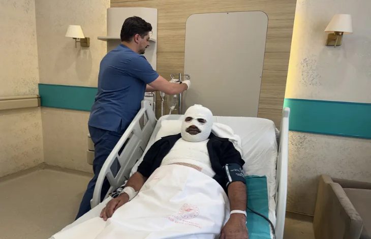 Görselde, hastane odasında tedavi gören bir hasta hastane yatağında uzanıyor. Hastanın başı ve vücudunun büyük bir bölümü bandajlarla sarılıyken, yanında bir sağlık çalışanı serum ekipmanıyla ilgileniyor. Oda, tıbbi ekipmanlar ve hasta yatağıyla donatılmış bir hastane ortamını yansıtıyor.