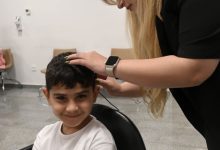 Görselde, kısa siyah saçlı ve beyaz tişörtlü küçük bir çocuk kameraya gülümseyerek bakarken, yanında bulunan bir uzman çocuğun kulağına ve başına bir cihaz yerleştiriyor veya ayarlama yapıyor. İşlem klinik ya da hastane ortamında gerçekleştiriliyor.