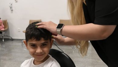 Görselde, kısa siyah saçlı ve beyaz tişörtlü küçük bir çocuk kameraya gülümseyerek bakarken, yanında bulunan bir uzman çocuğun kulağına ve başına bir cihaz yerleştiriyor veya ayarlama yapıyor. İşlem klinik ya da hastane ortamında gerçekleştiriliyor.