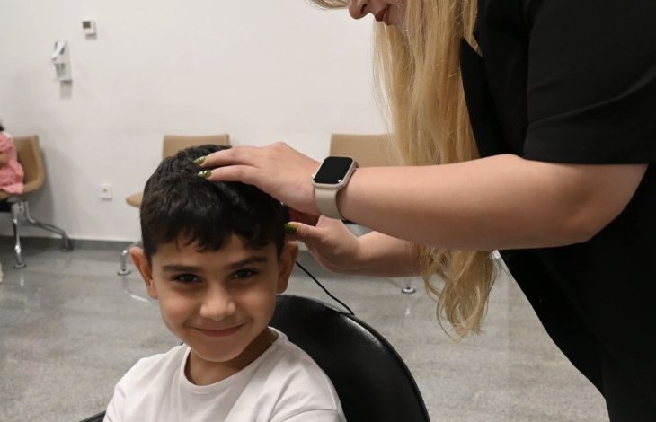 Görselde, kısa siyah saçlı ve beyaz tişörtlü küçük bir çocuk kameraya gülümseyerek bakarken, yanında bulunan bir uzman çocuğun kulağına ve başına bir cihaz yerleştiriyor veya ayarlama yapıyor. İşlem klinik ya da hastane ortamında gerçekleştiriliyor.