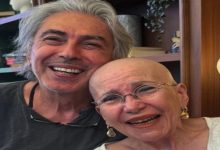 Görselde, başı tıraşlı ve gözlüklü yaşlı bir kadın ile yanında gri saçlı bir erkek samimi şekilde kameraya gülümseyerek poz veriyor. İkili birbirine yaslanmış durumda olurken, arka planda ahşap raflar ve dekoratif eşyalar bulunan sıcak bir iç mekân görülüyor.