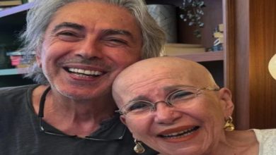 Görselde, başı tıraşlı ve gözlüklü yaşlı bir kadın ile yanında gri saçlı bir erkek samimi şekilde kameraya gülümseyerek poz veriyor. İkili birbirine yaslanmış durumda olurken, arka planda ahşap raflar ve dekoratif eşyalar bulunan sıcak bir iç mekân görülüyor.