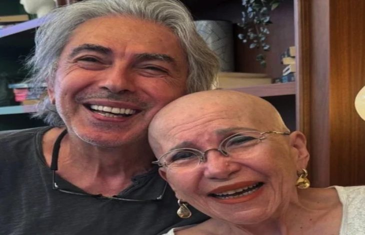 Görselde, başı tıraşlı ve gözlüklü yaşlı bir kadın ile yanında gri saçlı bir erkek samimi şekilde kameraya gülümseyerek poz veriyor. İkili birbirine yaslanmış durumda olurken, arka planda ahşap raflar ve dekoratif eşyalar bulunan sıcak bir iç mekân görülüyor.