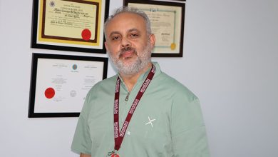 Prof. Dr. Ünal Aydın, iç mekânda, duvarda asılı çok sayıda diploma ve sertifikanın önünde duruyor. Üzerinde açık yeşil ameliyat önlüğü (scrubs) bulunuyor. Boynunda bordo renkte, üzerinde “Bezmialem Vakıf Üniversitesi” yazan bir yaka kartı askısı var. Arka plandaki çerçeveler; Sağlık Bakanlığı onaylı belgeler ve profesyonel sertifikalar gibi görünüyor. Aydın, duruşu ve ortamın düzeniyle klinik bir çalışma alanında, deneyimli bir cerrah izlenimi veriyor.