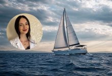 Duygu Yılmaz, uzun koyu saçları ve sakin gülümsemesiyle güven veren bir görünüme sahip. Beyaz bir gömlek ve zarif bir kolye takıyor; arka planda sade, açık tonlu bir duvar bulunuyor. Fotoğrafın arka fonunda, denizin ortasında ilerleyen küçük bir yelkenli yer alıyor. Hafif dalgalı suyun üzerinde beyaz yelkeniyle sakin bir yolculuk hissi veriyor. Gökyüzü parçalı bulutlu; ışık yelkenliye yumuşak bir parlaklık katıyor. Genel atmosfer; dinginlik, huzur ve farkındalık temasını destekleyen sade bir uyum taşıyor.