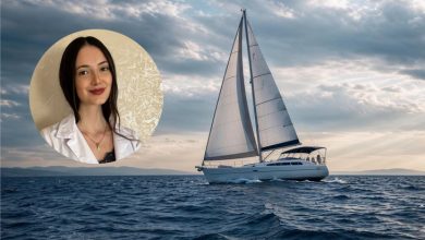 Duygu Yılmaz, uzun koyu saçları ve sakin gülümsemesiyle güven veren bir görünüme sahip. Beyaz bir gömlek ve zarif bir kolye takıyor; arka planda sade, açık tonlu bir duvar bulunuyor. Fotoğrafın arka fonunda, denizin ortasında ilerleyen küçük bir yelkenli yer alıyor. Hafif dalgalı suyun üzerinde beyaz yelkeniyle sakin bir yolculuk hissi veriyor. Gökyüzü parçalı bulutlu; ışık yelkenliye yumuşak bir parlaklık katıyor. Genel atmosfer; dinginlik, huzur ve farkındalık temasını destekleyen sade bir uyum taşıyor.