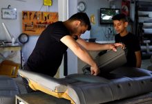Görselde bir oto döşeme atölyesinde çalışan genç bir kişi, araç koltuğunun döşemesini onarırken görülüyor. Yanında duran bir çocuk çalışmayı izlerken, arka planda tamir ekipmanları ve atölye malzemeleri yer alıyor.