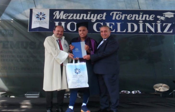 Otizmli Milli Kayakçı Muhsin Murat Bingül'e tören sahnesinde, sağında ve solunda bulunan yetkili erkekler diplomasını takdim ediyor.