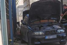 Görselde, kaputu açık siyah bir otomobil dar bir alanda park halinde duruyor. Aracın motor bölmesi açıkken sağ tarafta bir kişi otomobilin yanında ayakta bulunuyor. Çevresi duvarlarla çevrili alanda bakım veya inceleme yapıldığı izlenimi oluşuyor.