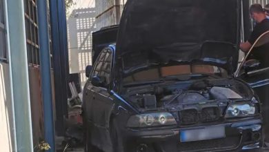 Görselde, kaputu açık siyah bir otomobil dar bir alanda park halinde duruyor. Aracın motor bölmesi açıkken sağ tarafta bir kişi otomobilin yanında ayakta bulunuyor. Çevresi duvarlarla çevrili alanda bakım veya inceleme yapıldığı izlenimi oluşuyor.