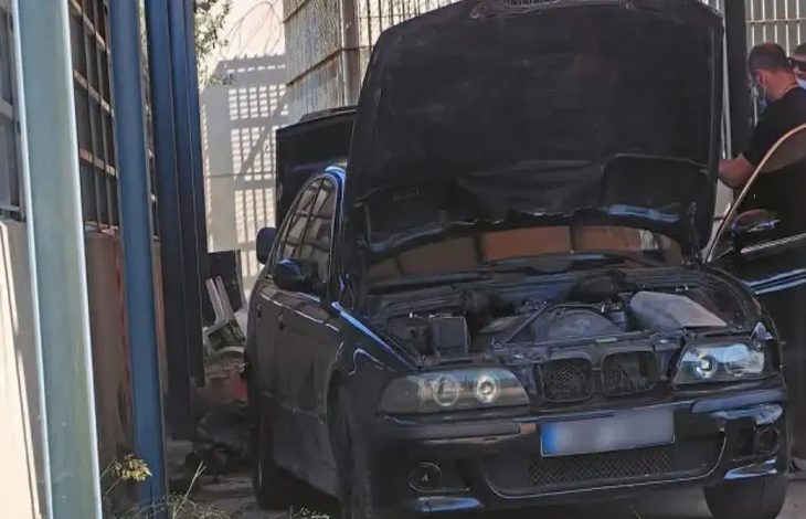 Görselde, kaputu açık siyah bir otomobil dar bir alanda park halinde duruyor. Aracın motor bölmesi açıkken sağ tarafta bir kişi otomobilin yanında ayakta bulunuyor. Çevresi duvarlarla çevrili alanda bakım veya inceleme yapıldığı izlenimi oluşuyor.