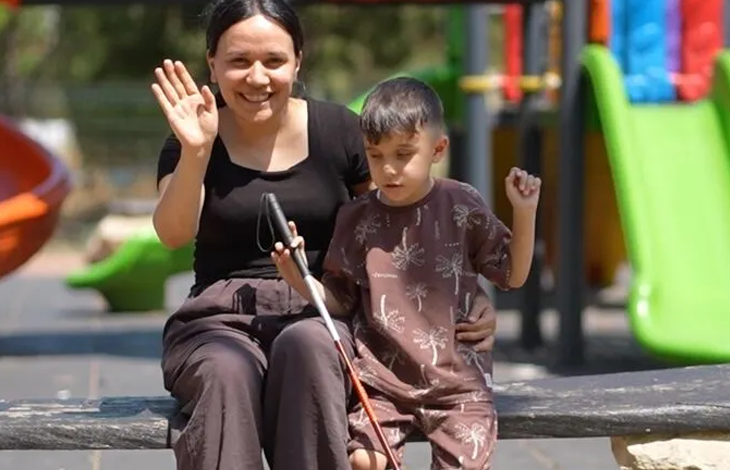 Görme engelli Toprak, oyun parkında annesinin yanında beyaz bastonunu tutarak ayakta duruyor.