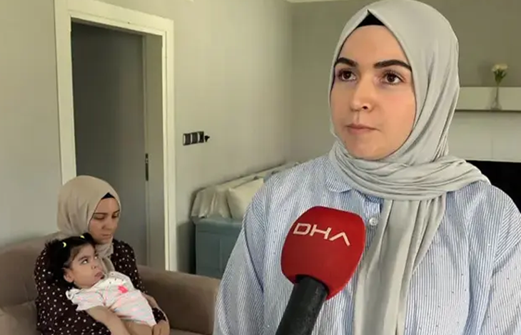 Görselde, açık renk başörtülü bir kadın DHA mikrofonuna konuşuyor. Arka planda koltukta oturan başka bir kadın, kucağında bir bebeği tutuyor.