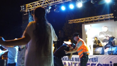 Görselde gece düzenlenen açık hava etkinliğinde sahne önünde bulunan bir çocuk, turuncu yelekli zabıta görevlisi tarafından pistten uzaklaştırılmaya çalışılıyor. Arka planda sahnede müzisyenler ve etkinliği izleyen kişiler görülüyor.